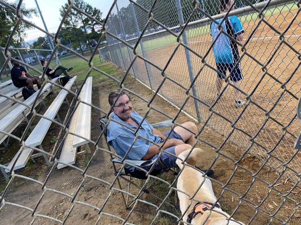 Dave sitting in a chair at a baseball field cheering on his Hail Mary friends and family.  